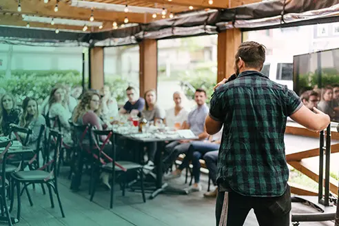Male speaker in a black and green flannel shirt giving a speech to an audience at a restaurant.